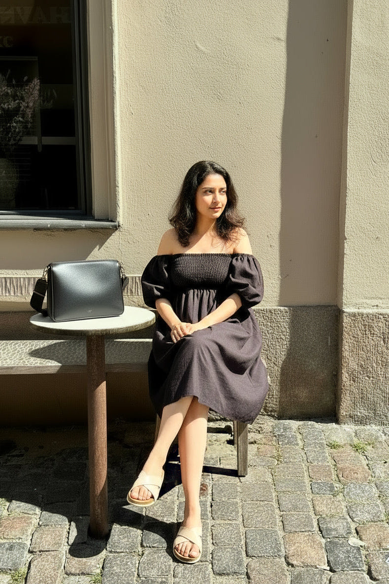Woman in a gray off-shoulder dress sitting on a chair next to a table with a bag on a stone pavement.