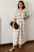 A woman stands indoors wearing a loose white midi dress with a yellow and orange floral print, holding a dark brown clutch bag. She is standing against a plain off-white wall on a wooden floor.