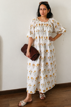 A woman stands indoors wearing a loose white midi dress with a yellow and orange floral print, holding a dark brown clutch bag. She is standing against a plain off-white wall on a wooden floor.