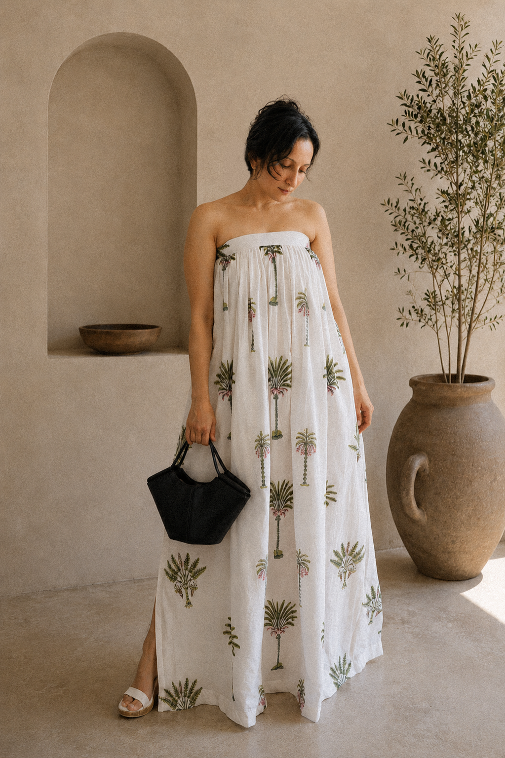 Woman wearing a strapless dress with palm tree patterns, showcasing the side slit and front look. The back has elastic band and a tie for comfort of wearing in a warm-toned indoor setting.