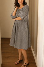Woman wearing a navy and white checkered dress standing in a room with wooden floor and beige walls.