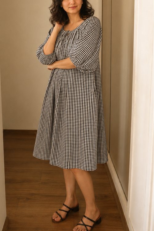 Woman wearing a black and white checkered dress standing in a room with wooden floor and beige walls.