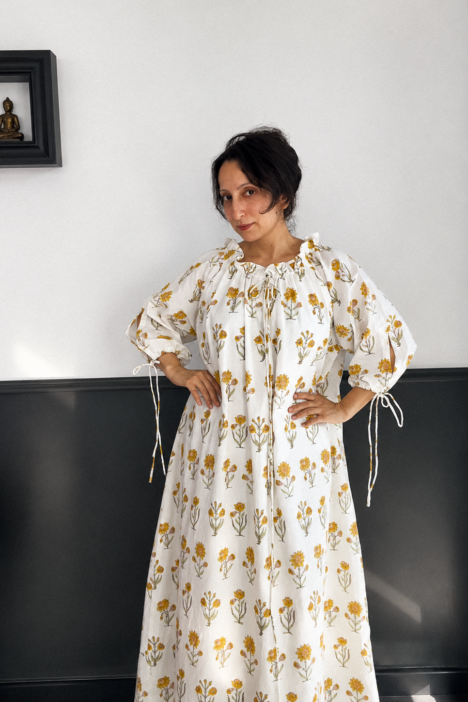 Woman wearing a floral dress with cream base and small yellow flower all over. dress has neck tie and ties around the cuffs for ease of wearing. 
