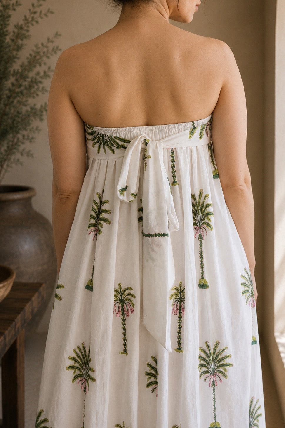 Woman wearing a strapless dress with palm tree patterns, showcasing the back that has elastic band and a tie for comfort of wearing in a warm-toned indoor setting.