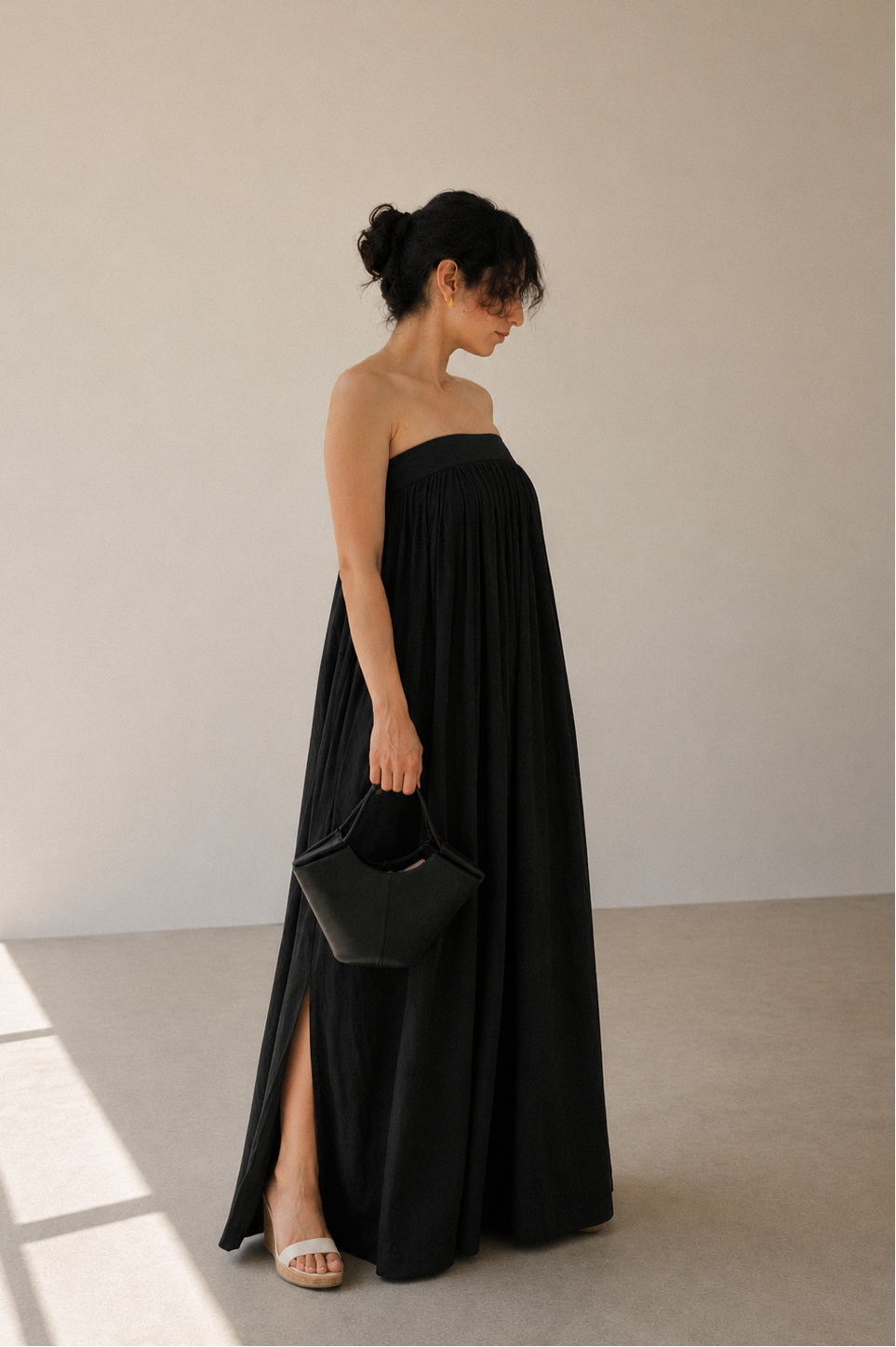 Woman in balmy nights black strapless maxi dress with a side slit, holding a black handbag against a plain background