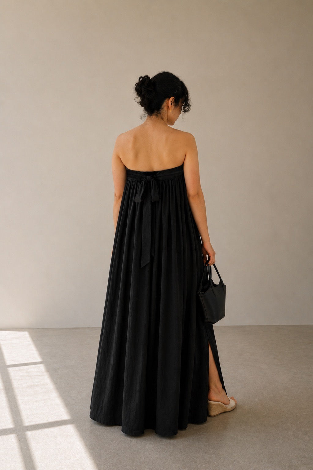 Woman wearing balmy nights black cotton strapless maxi dress with back ties, holding a small black handbag against a plain background.