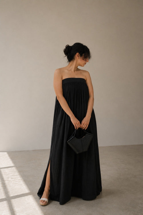Woman wearing balmy nights black strapless maxi dress with side slit holding a black handbag against a plain background