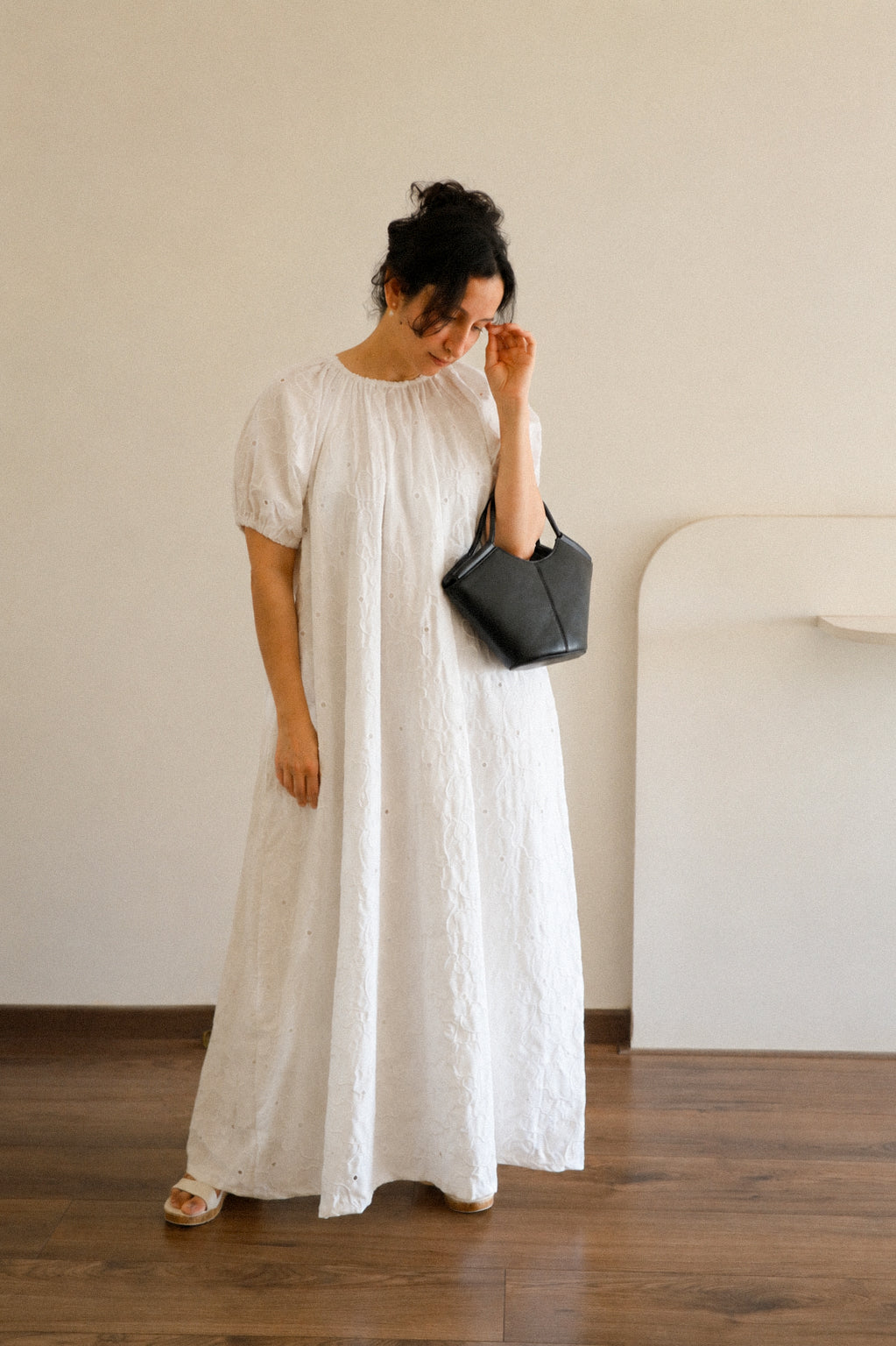Woman in a Cloudline White Eyelet Cotton Maxi Dress With Puff Sleeve holding a black handbag in a minimalistic room.
