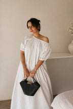Woman in a Cloudline White Eyelet Cotton Maxi Dress With Puff Sleeve, wearing it Off-Shoulder style, holding a black handbag in a minimalistic room.