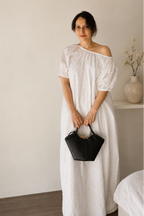 Woman in a Cloudline White Eyelet Cotton Maxi Dress With Puff Sleeve, wearing it Off-Shoulder style, holding a black handbag in a minimalistic room.