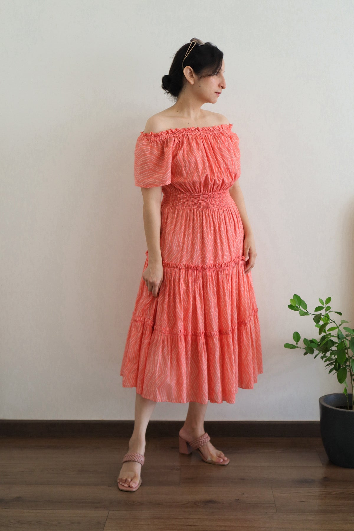 Woman wearing Coral Peach Mulmul Cotton Tiered Midi Dress off-shoulder standing against a plain wall with a plant in the background.