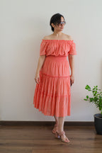 Woman wearing Coral Peach Mulmul Cotton Tiered Midi Dress off-shoulder standing against a plain wall with a plant in the background.