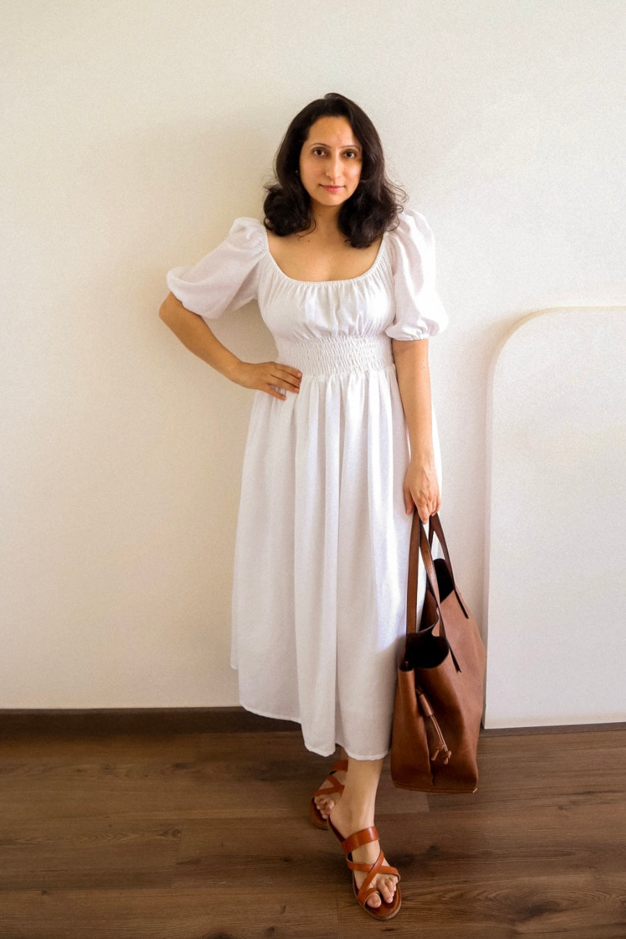 Woman in Dawn White Cotton Smocked Waist Midi Dress holding a brown bag against a plain wall.