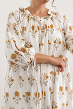 Close up of woman wearing a floral dress with cream base and small yellow flower all over. She shows the close up of dress that has neck tie and ties around the cuffs for ease of wearing. 