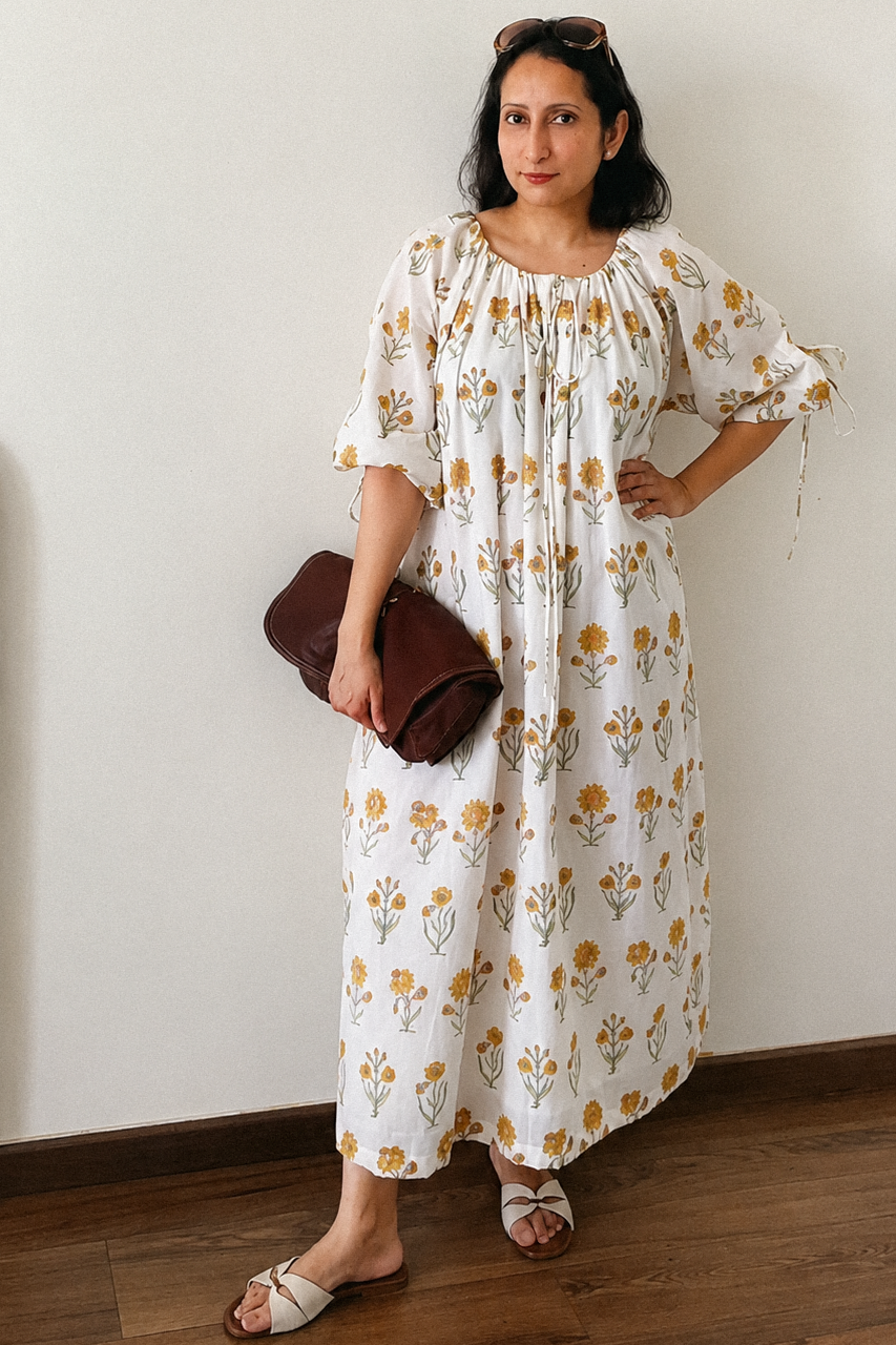 A woman stands indoors wearing a loose white midi dress with a yellow and orange floral print, holding a dark brown clutch bag. She is standing against a plain off-white wall on a wooden floor.