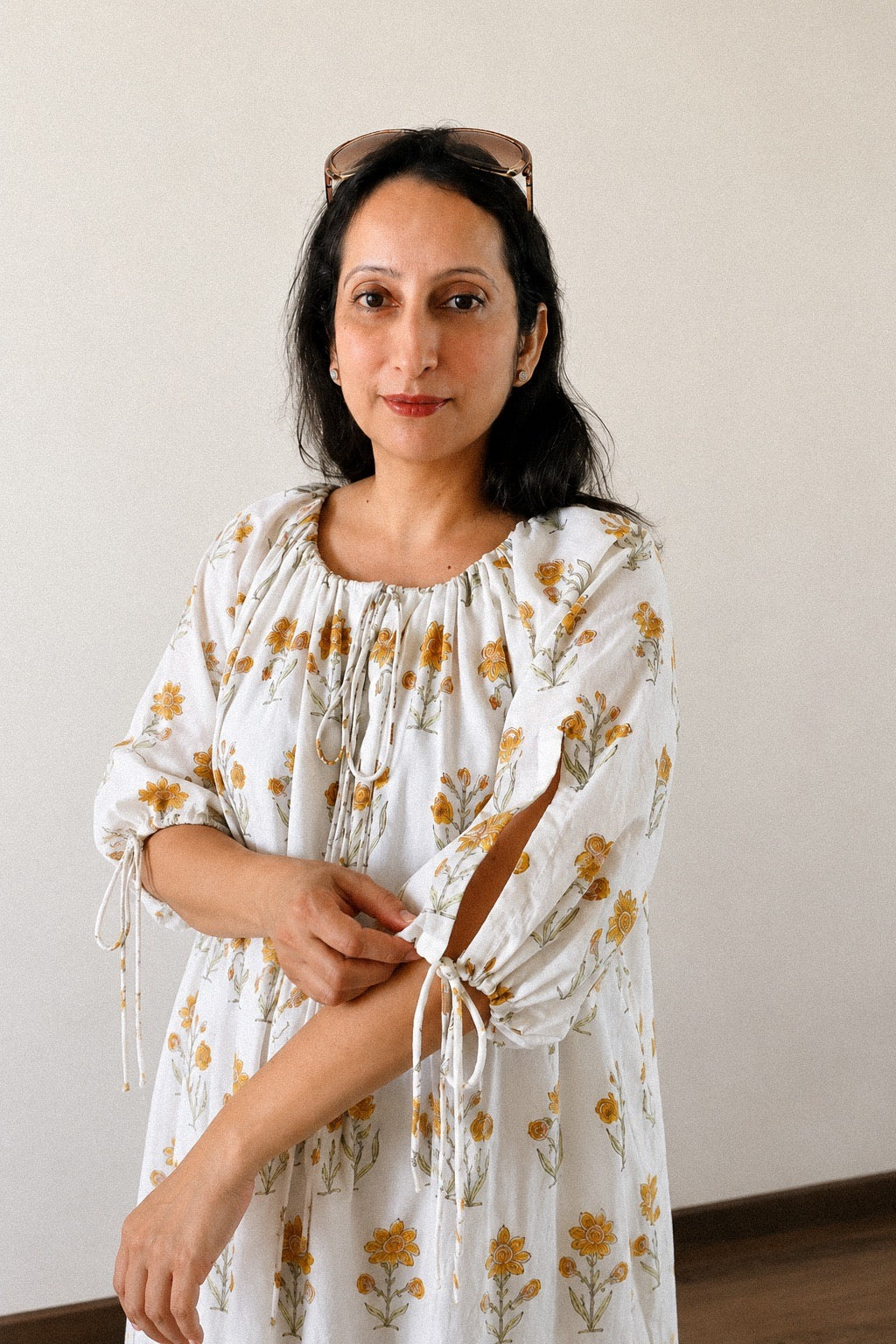 A woman wearing a gathered neck, long-tie-sleeve dress with a yellow and green floral print, standing against a plain off-white wall.