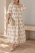 A woman models Daydream Cream Floral midi dress, cream-colored, long-sleeved midi dress with a yellow floral pattern, styled with a beige clutch and neutral sandals in a sunlit setting.