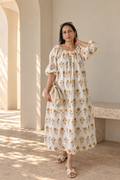 A woman models daydream cream floral midi dress, cream-colored, long-sleeved midi dress with a yellow floral pattern, styled with a beige clutch and neutral sandals in a sunlit setting.