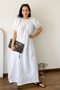 A woman wearing a white eyelet puff-sleeve maxi dress, holding a black and white patterned clutch, standing in a minimalist room with a beige wall and wooden floor.