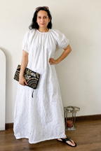 A woman wearing a white eyelet puff-sleeve maxi dress, holding a black and white patterned clutch, standing in a minimalist room with a beige wall and wooden floor.