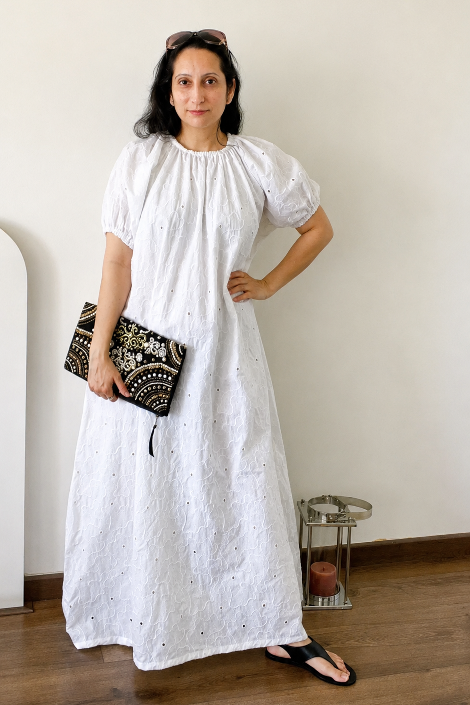 A woman wearing a white eyelet puff-sleeve maxi dress, holding a black and white patterned clutch, standing in a minimalist room with a beige wall and wooden floor.