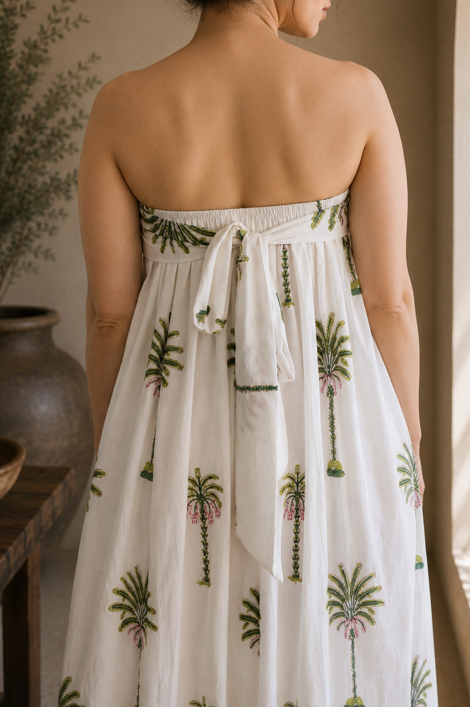 Woman wearing a strapless dress with palm tree patterns, showcasing the back that has elastic band and a tie for comfort of wearing in a warm-toned indoor setting.