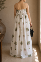 Woman wearing a strapless dress with palm tree patterns, showcasing the back that has elastic band and a tie for comfort of wearing in a warm-toned indoor setting.