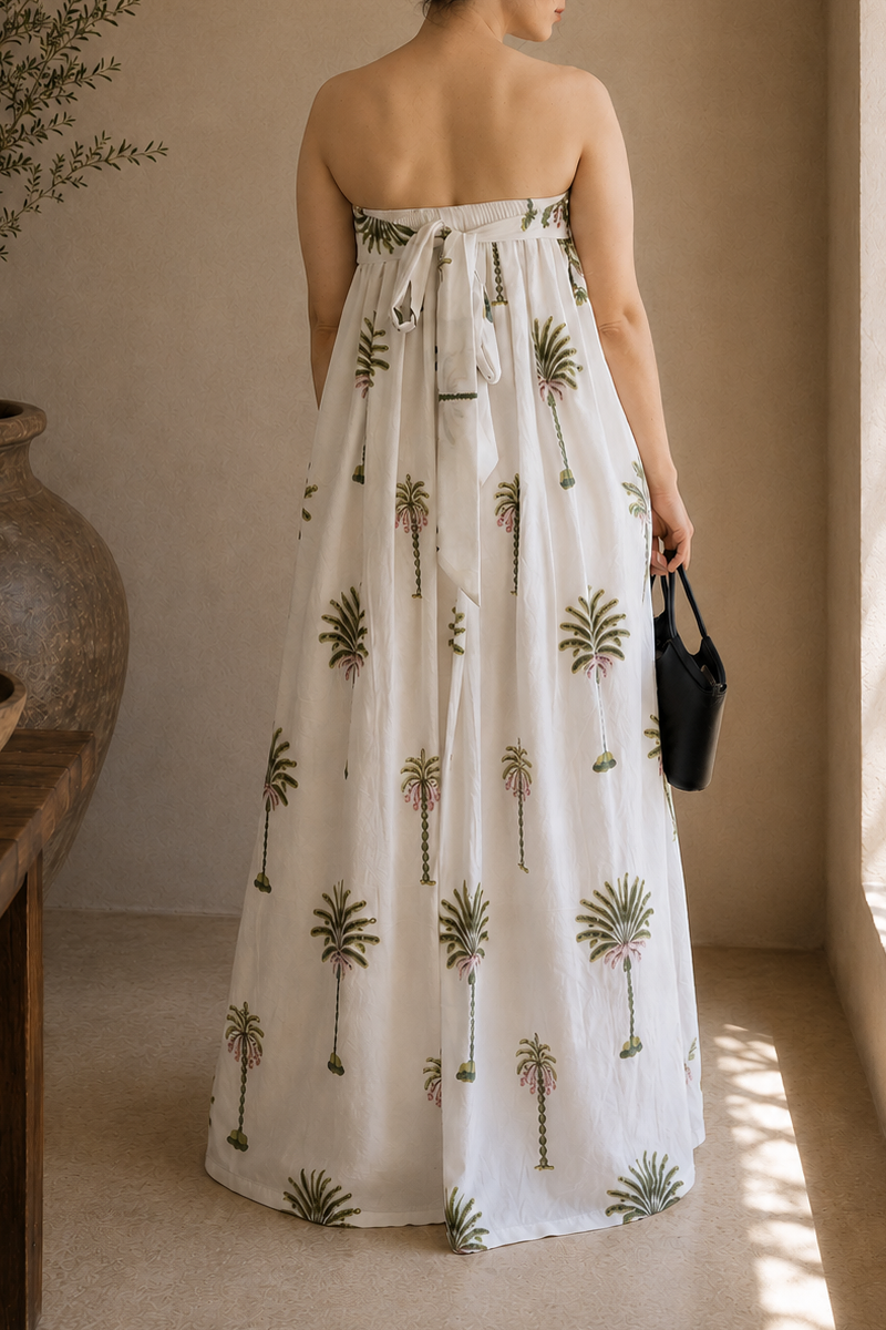 Woman wearing a strapless dress with palm tree patterns, showcasing the back that has elastic band and a tie for comfort of wearing in a warm-toned indoor setting.
