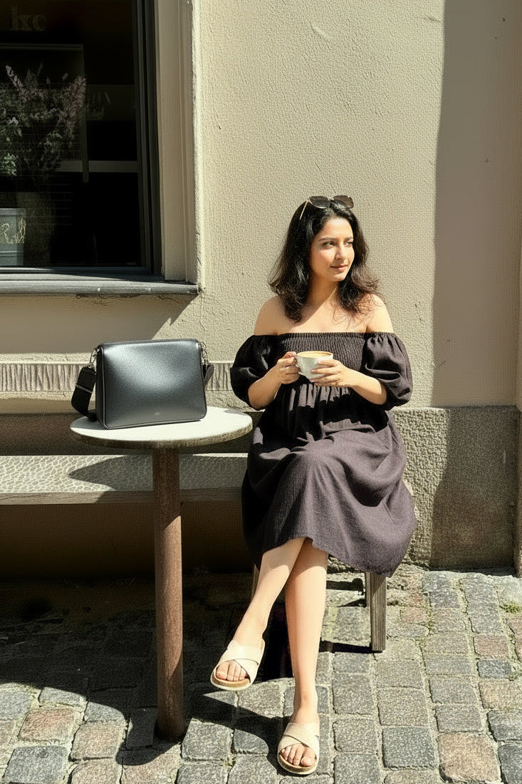 Woman in Palm dress in midnight black linen with shirred bodice and puff sleeves - front view, sitting on a chair holding a cup, with a handbag next to her on a small table, against a beige wall.