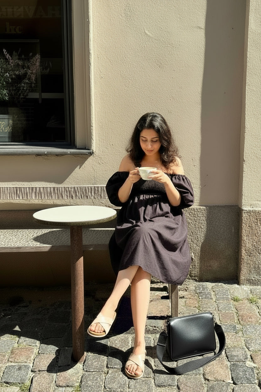 Woman in Palm dress in midnight black linen with shirred bodice and puff sleeves - front view, sitting on a chair holding a cup, with a handbag next to her on a small table, against a beige wall.