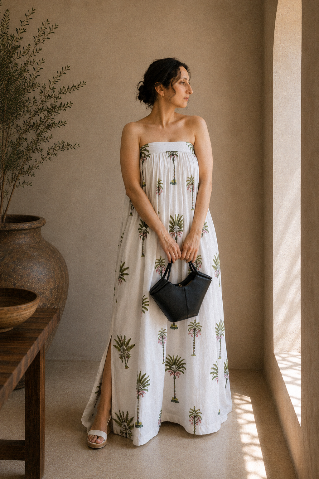 Woman wearing a strapless dress with palm tree patterns, showcasing the side slit and front look. The back has elastic band and a tie for comfort of wearing in a warm-toned indoor setting.