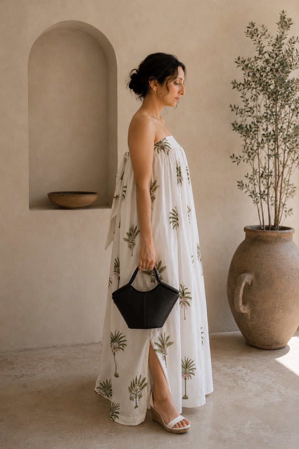 Woman wearing a strapless dress with palm tree patterns, showcasing the side that has a slit and back that has elastic band and a tie for comfort of wearing in a warm-toned indoor setting.