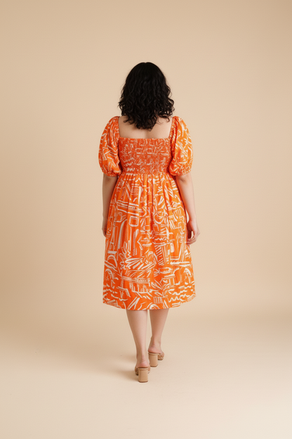 Woman wearing an orange patterned cotton midi dress with shirred bodice and puffed sleeves on a beige background