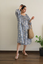 Woman in Still Blue Cotton Floral Midi Dress Puff Sleeve - Pastel Blue dress holding a woven bag indoors.
