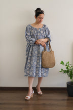 Woman in Still Blue Cotton Floral Midi Dress Puff Sleeve - Pastel Blue dress holding a woven bag indoors.