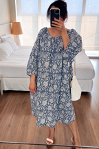 Woman taking a mirror selfie wearing Still Blue Cotton Floral Midi Dress Puff Sleeve - Pastel Blue dress holding a white sling bag in a bedroom.