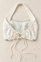 An off-white halter crop top with a tie-front closure, delicate lace trim at the neckline, and gray patterned cuffs, laid flat on a light beige background.