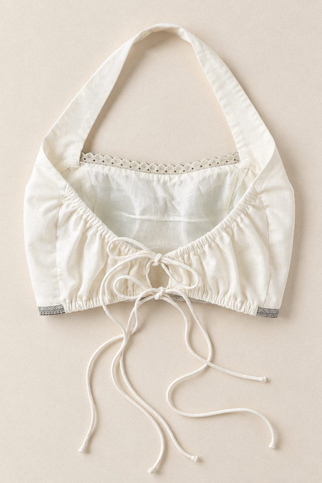 An off-white halter crop top with a tie-front closure, delicate lace trim at the neckline, and gray patterned cuffs, laid flat on a light beige background.